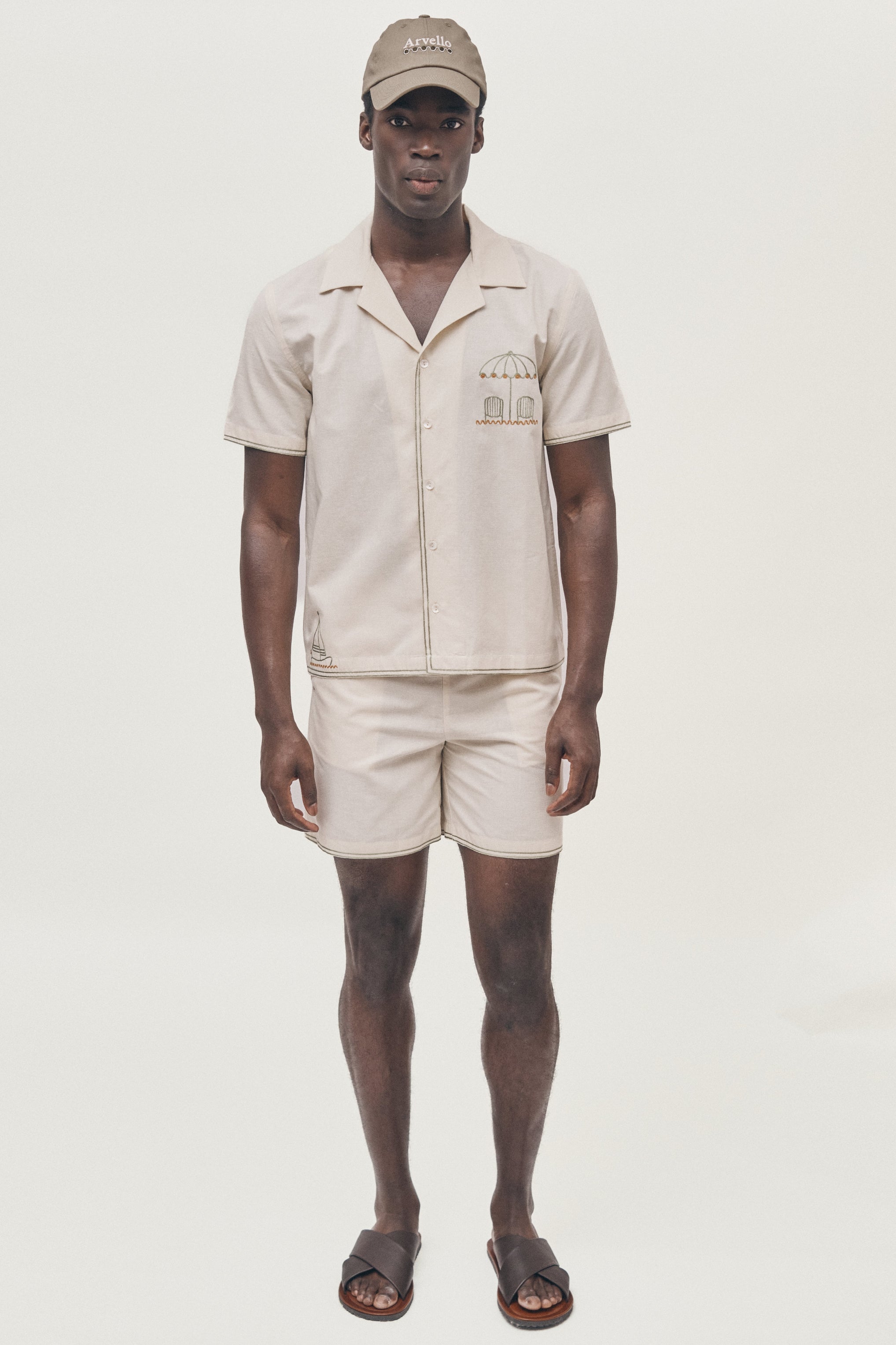 Man wearing a beige shirt and shorts set with a cap on a white background