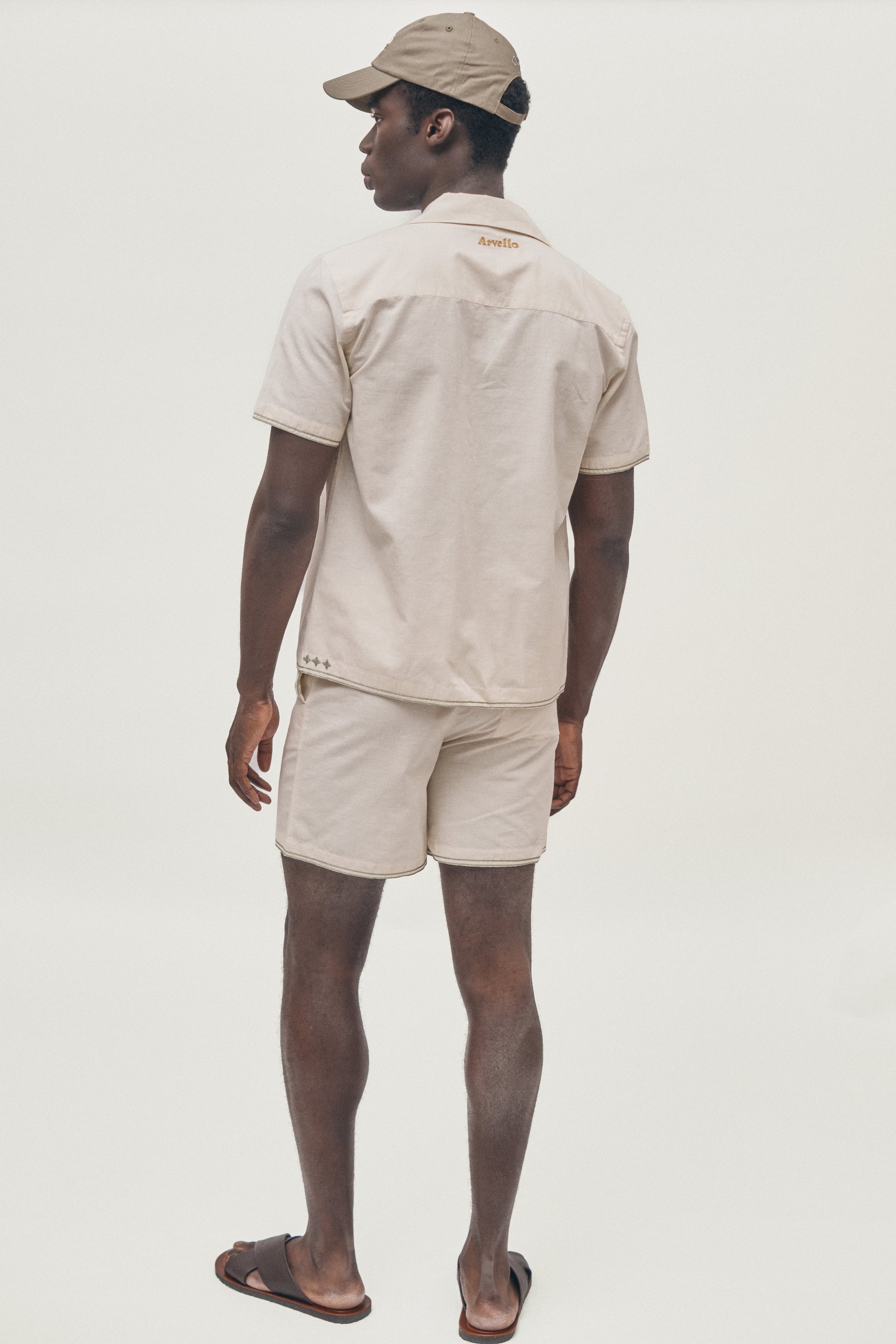 Man wearing a beige shirt and shorts set with a cap on a white background