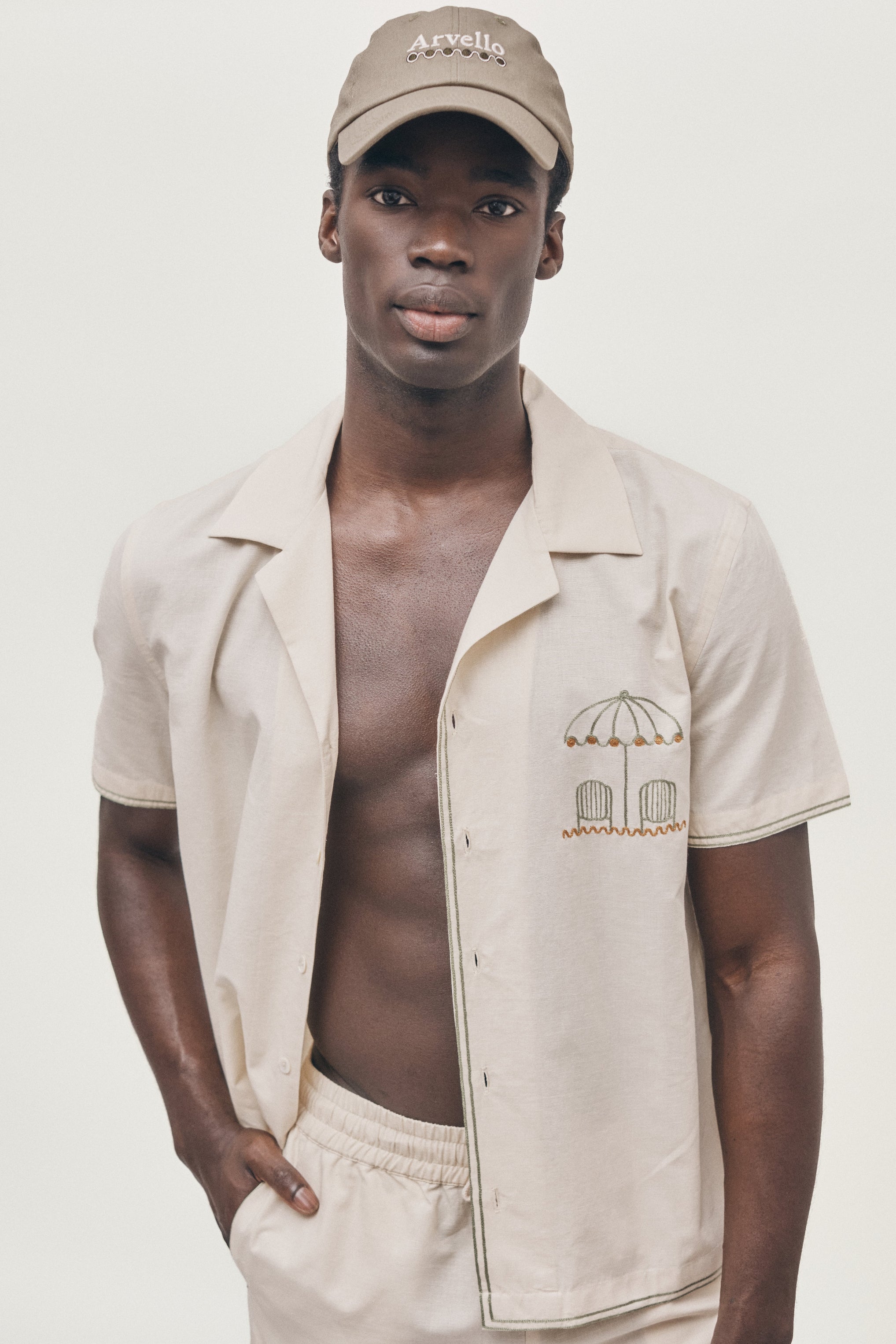 Man wearing a beige shirt with an embroidered design and cap on a plain background