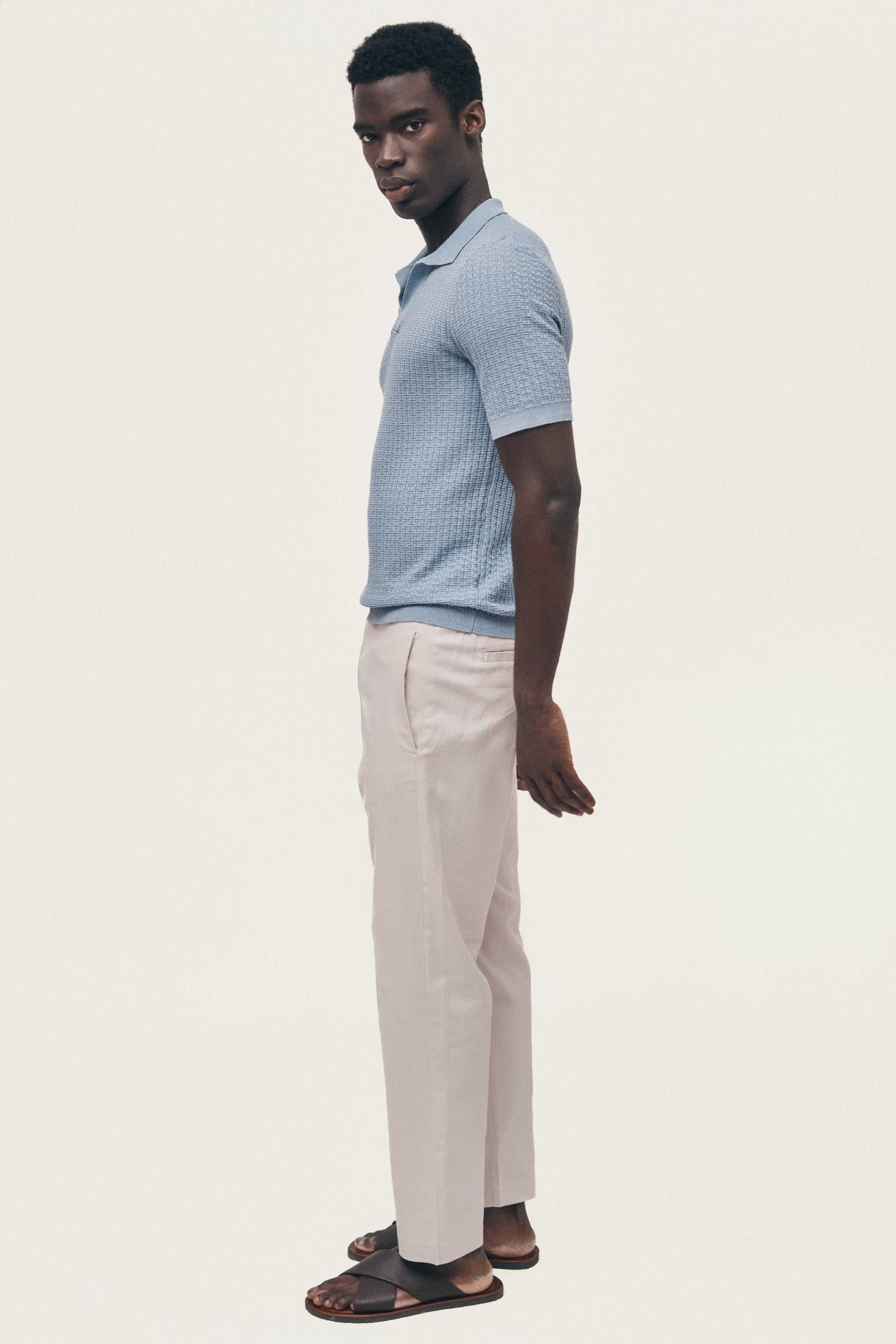 Man wearing a light blue patterned polo shirt and beige pants on a white background